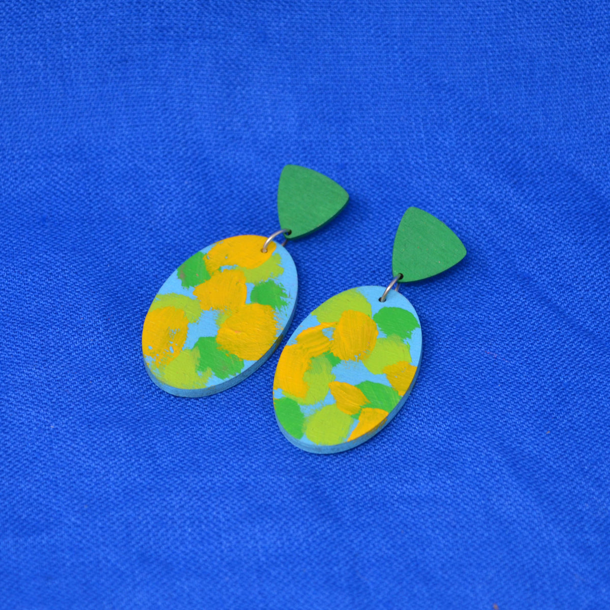 A pair of handmade earrings featuring oval drops with a textured yellow and green paint pattern on a light blue base, hanging from green triangle-shaped studs against a vibrant blue fabric background.