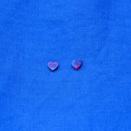 A pair of small heart-shaped stud earrings with a painted pattern of blue, purple, and red, displayed on a textured blue fabric background.