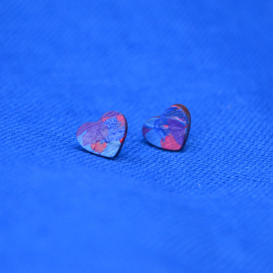 A pair of small heart-shaped stud earrings with a painted pattern of blue, purple, and red, displayed on a textured blue fabric background.