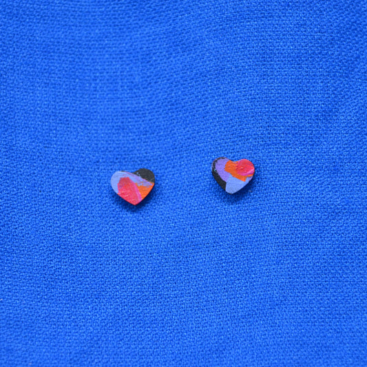 A pair of small heart-shaped stud earrings with a multicolor abstract pattern, displayed on a textured blue fabric background.