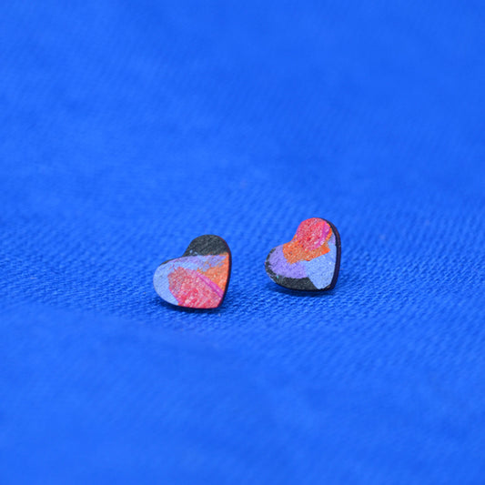 A pair of small heart-shaped stud earrings with a marbled, abstract pattern in pink, orange, purple, and black, displayed on a textured blue background.