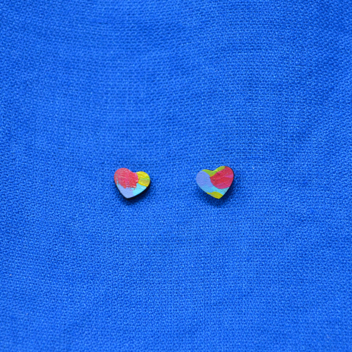 Two small heart-shaped stud earrings with a multicolor abstract paint-stroke pattern, displayed on a textured blue fabric background.