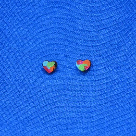 Two small heart-shaped stud earrings with a colorful marbled pattern in green, red, orange, and blue, displayed on a textured blue fabric background.