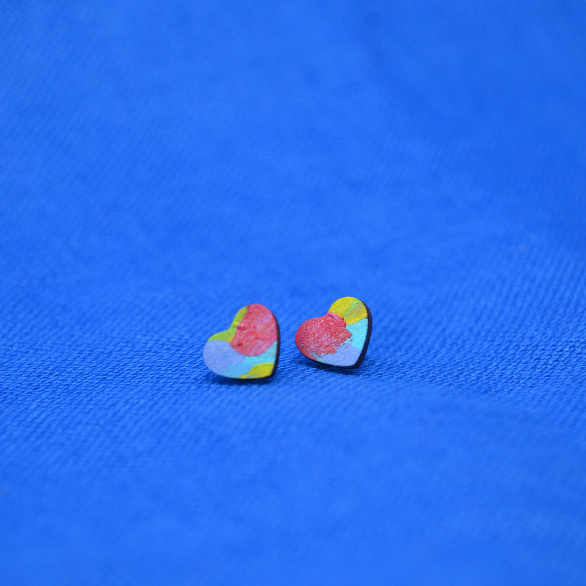 A pair of small heart-shaped stud earrings with a colorful, textured paint-splatter design in pink, yellow, blue, and green, resting on a textured blue background.