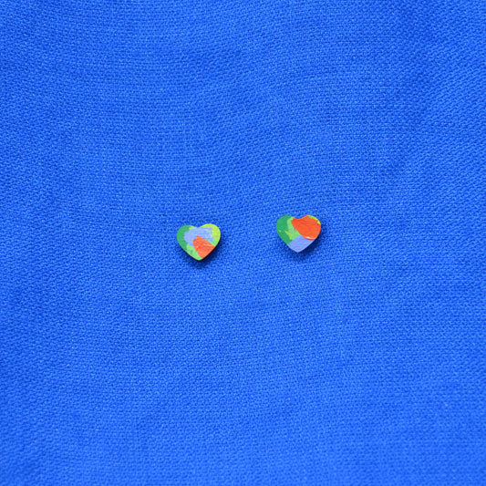 Two small wooden heart-shaped stud earrings with a multicolor painted pattern of green, red, orange, and yellow, displayed on a textured blue fabric background.