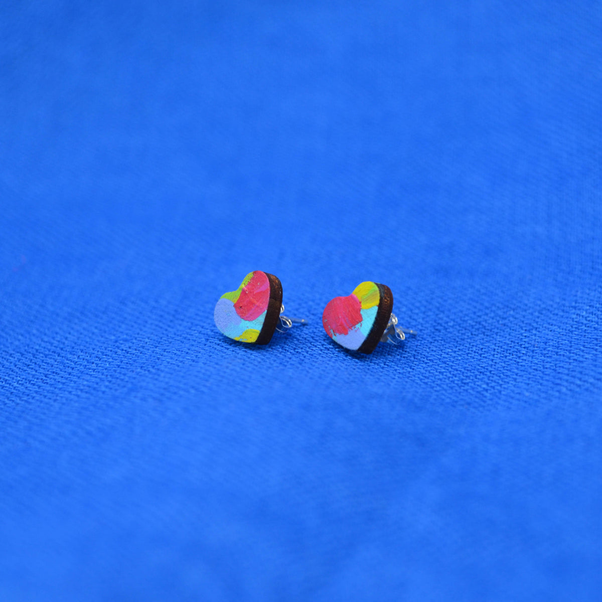 A pair of small heart-shaped stud earrings with a colorful abstract pattern on a textured blue background.