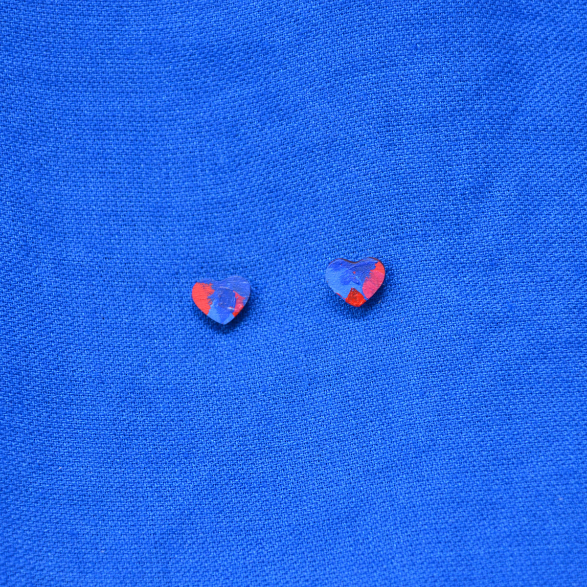 A pair of small heart-shaped earrings with a painted blue and red pattern, displayed on a textured blue background.