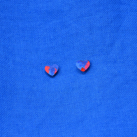 A pair of small heart-shaped earrings with a painted blue and red pattern, displayed on a textured blue background.