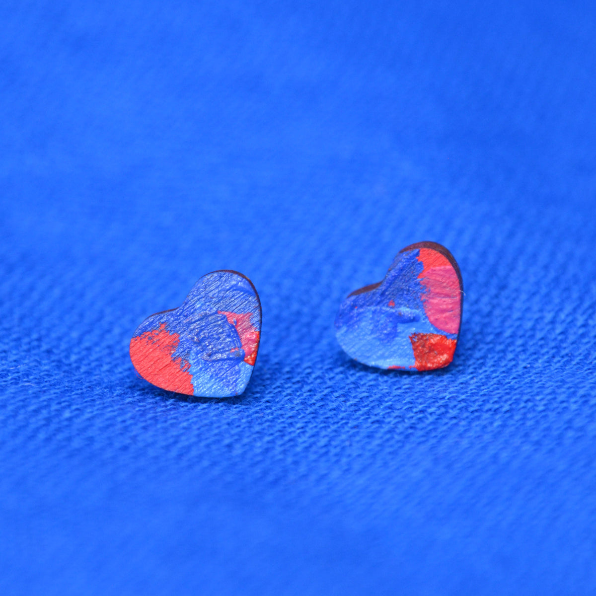 A pair of small heart-shaped earrings with a painted blue and red pattern, displayed on a textured blue background.
