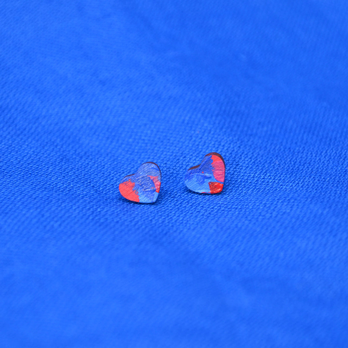 A pair of small heart-shaped earrings with a painted blue and red pattern, displayed on a textured blue background.