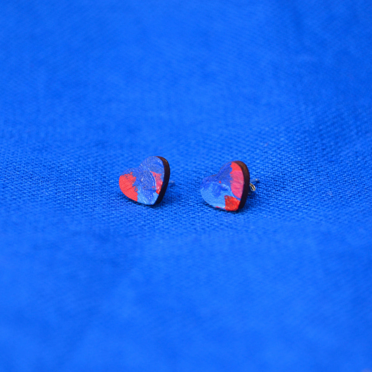 A pair of small heart-shaped stud earrings with a blue and red marbled pattern and wooden edges, displayed on a textured bright blue fabric background.