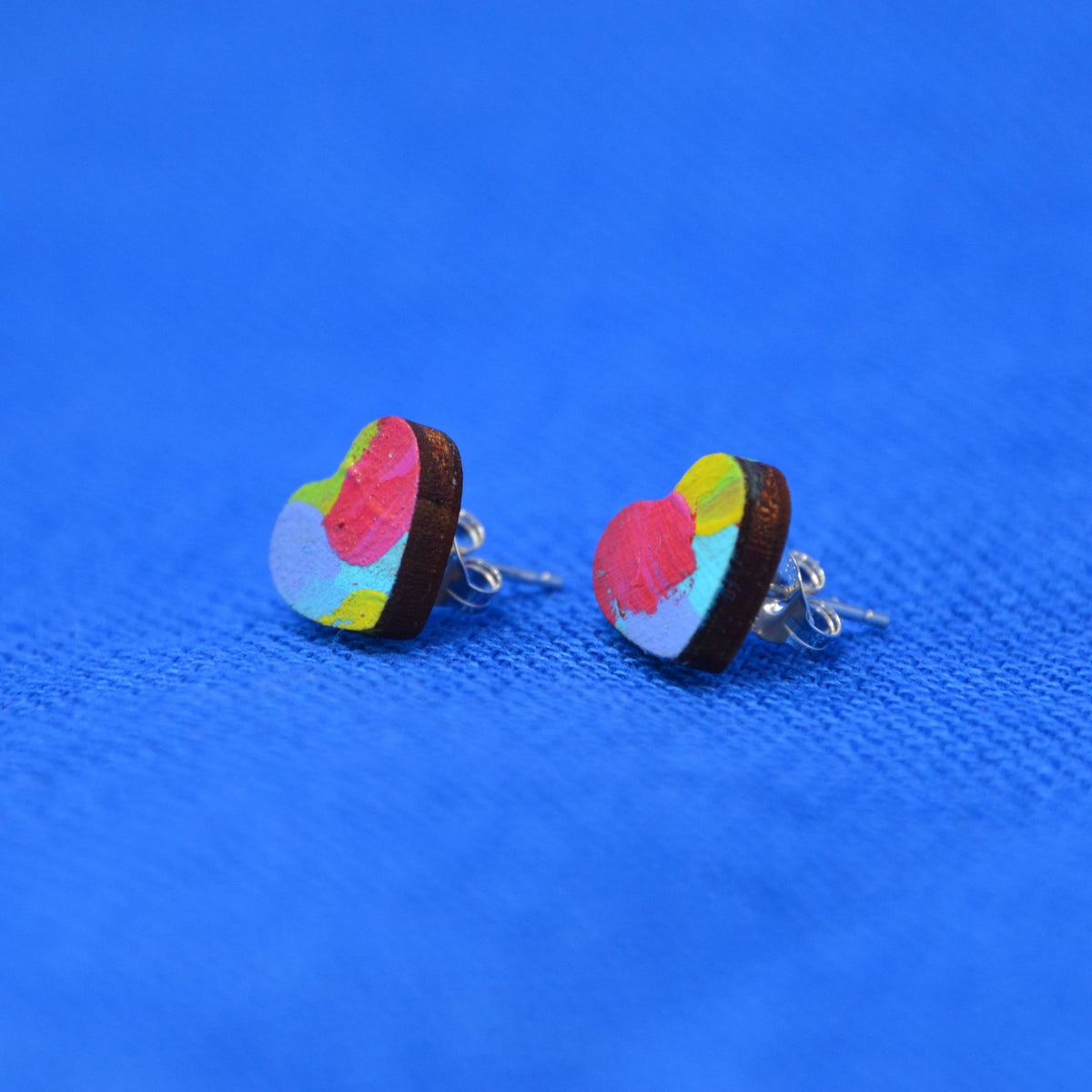 A pair of heart-shaped stud earrings with a colorful abstract paint-stroke pattern on a dark brown wooden base, displayed on a textured blue background.