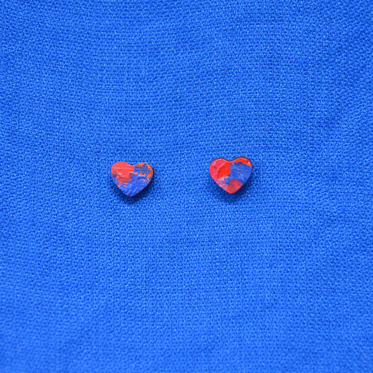 Two small heart-shaped stud earrings with a painted red and blue pattern, displayed on a textured blue fabric background.