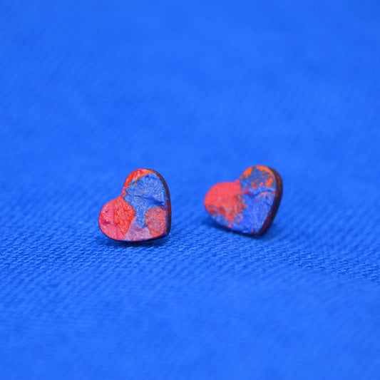 A pair of small heart-shaped stud earrings with a marbled red and blue pattern and wooden edges, displayed on a textured blue background.