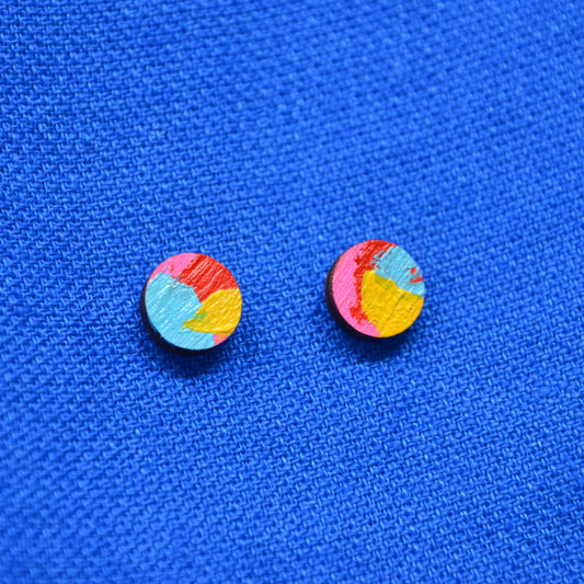 A pair of small round stud earrings with a colorful abstract painted design in pink, red, yellow, and light blue, displayed on a textured blue fabric background.
