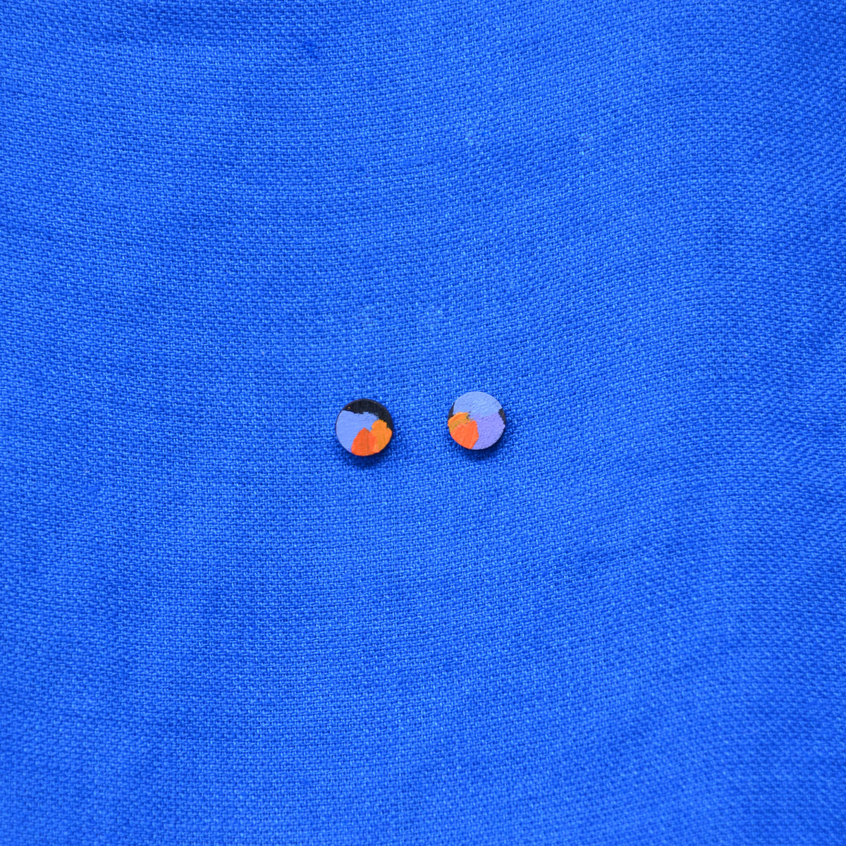 A pair of small wooden round stud earrings with a black circular base and a colorful abstract design in orange and purple, displayed on a textured blue background.