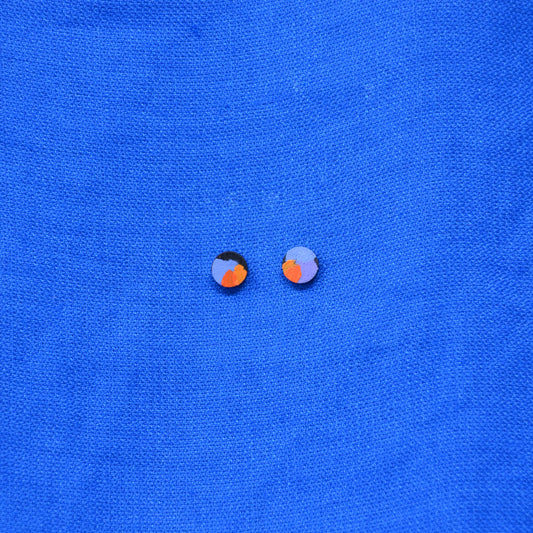 A pair of small wooden round stud earrings with a black circular base and a colorful abstract design in orange and purple, displayed on a textured blue background.