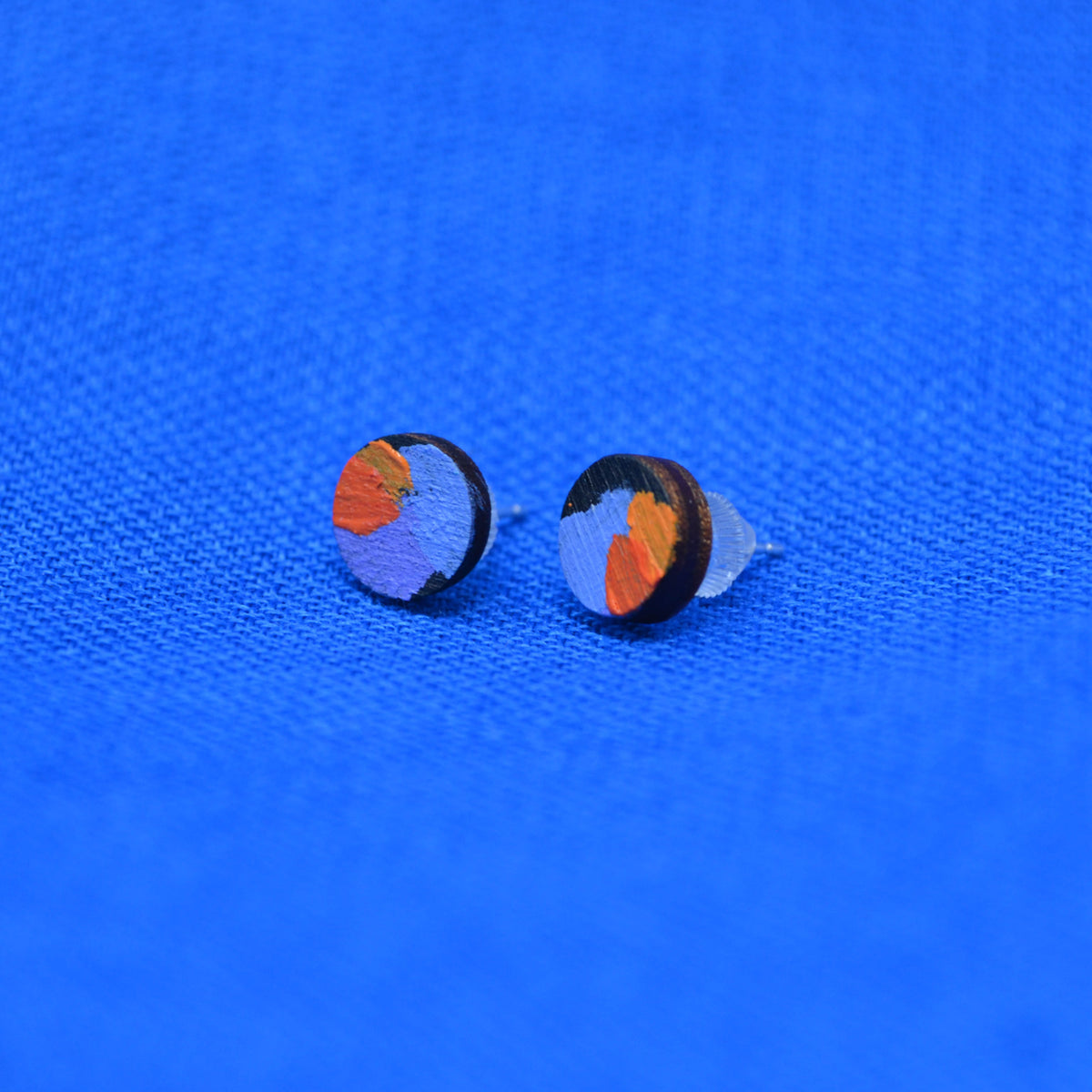 A pair of round stud earrings with a colorful abstract design in orange, purple, and black, displayed on a textured blue fabric background.