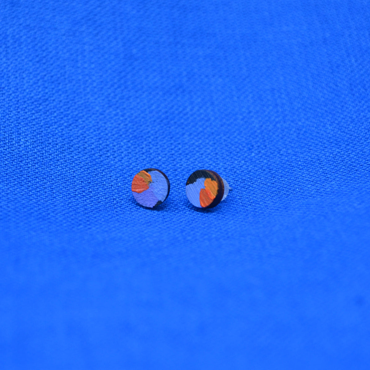 A pair of small wooden round stud earrings with a black circular base and a colorful abstract design in orange and purple, displayed on a textured blue background.
