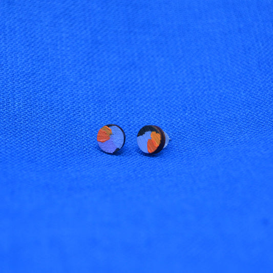 A pair of small wooden round stud earrings with a black circular base and a colorful abstract design in orange and purple, displayed on a textured blue background.