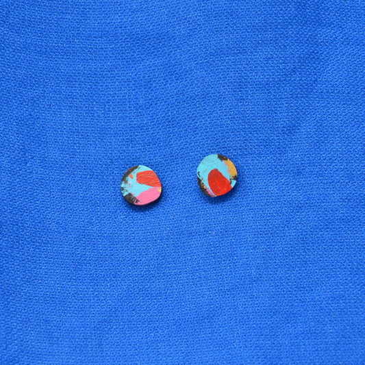 A pair of wooden round stud earrings with a colorful abstract pattern of turquoise, red, pink, and black with a hint of gold, displayed on a textured blue background.