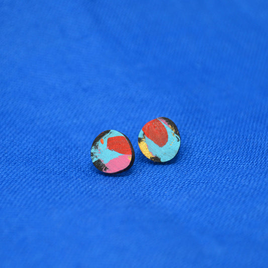 A pair of wooden round stud earrings with a colorful abstract pattern of turquoise, red, pink, and black with a hint of gold, displayed on a textured blue background.