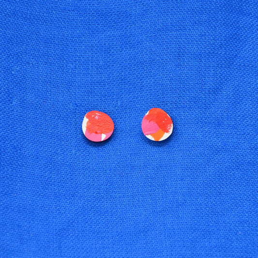 A pair of blob round stud earrings with a painted abstract red, white, pink and orange pattern on a wooden base, displayed on a vibrant blue textured fabric background.