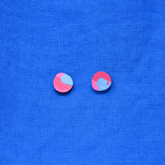Two small wooden oval stud earrings with a pink, light blue, and dark painted pattern, displayed on a textured blue fabric background.