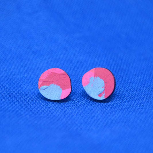 Two small wooden oval stud earrings with a pink, light blue, and dark painted pattern, displayed on a textured blue fabric background.