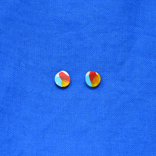A pair of small round stud earrings with a colorful marbled pattern in red, yellow, light blue, and white, displayed on a textured blue fabric background.