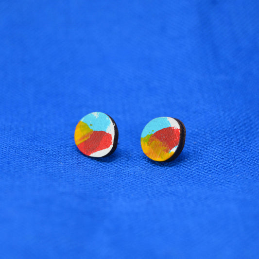 A pair of round stud earrings with abstract multicolor painted designs in blue, red, yellow, and white on a wooden base, displayed on a textured blue background.
