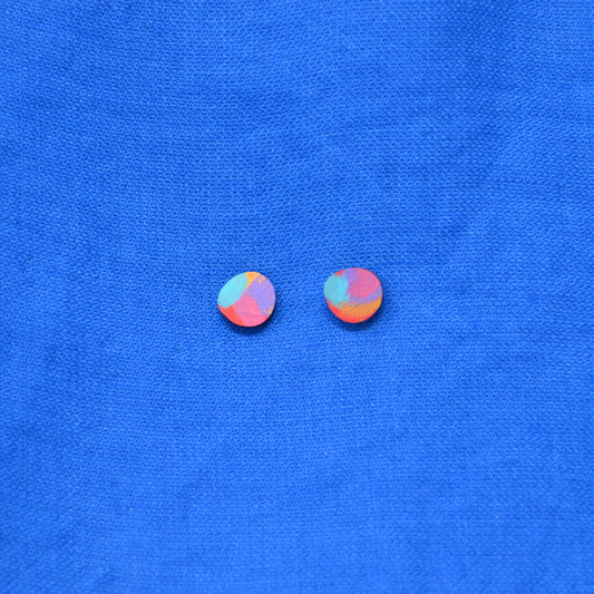 A pair of small multicolor blob circle stud earrings with layered paint in gold, pink, purple, red and blue, displayed on a textured blue background.