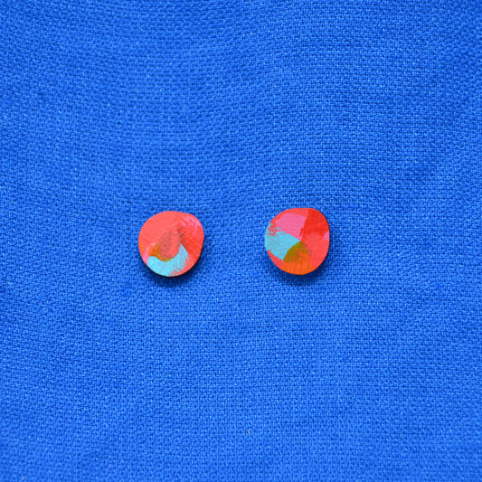 A pair of small multicolor blob circle stud earrings with layered paint in green, pink,  red and blue, displayed on a textured blue background.