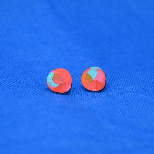A pair of round, flat stud earrings with a textured, abstract red and turquoise paint design, displayed on a textured blue background.