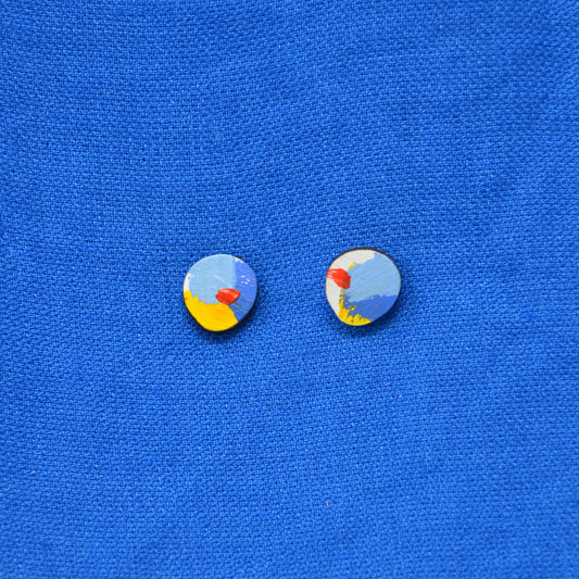 A pair of wooden small oval stud earrings with a colorful abstract design featuring light blue, yellow, red, and white, displayed on a textured blue background.