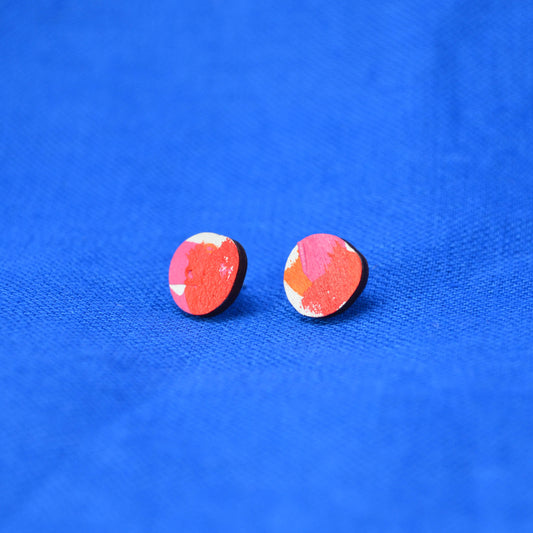 A pair of blob round stud earrings with a painted abstract red, white, pink and orange pattern on a wooden base, displayed on a vibrant blue textured fabric background.