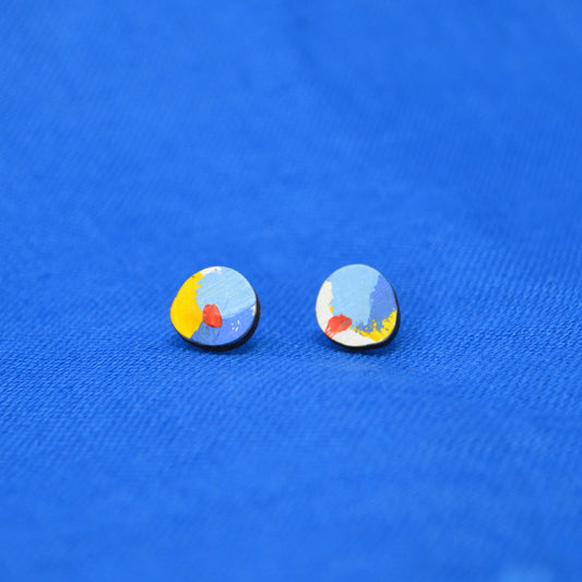 A pair of small oval stud earrings with a colorful abstract design featuring light blue, yellow, red, and white, displayed on a textured blue background.