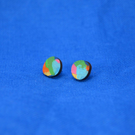 A pair of small round stud earrings with a multicolor abstract pattern of green, blue, pink, and orange on a wooden backing, displayed on a textured blue fabric background.