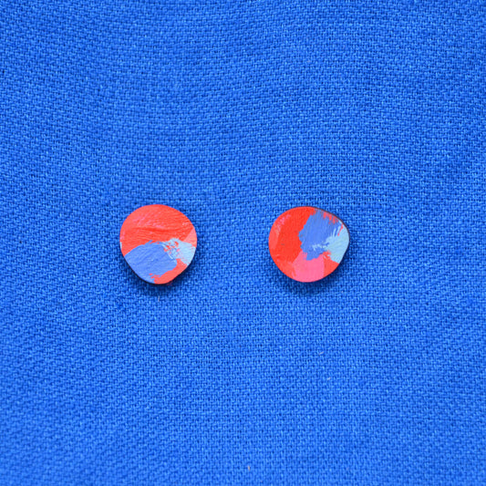 A pair of wooden blob round stud earrings with a red, blue, and pink abstract painted design, displayed on a textured blue background.