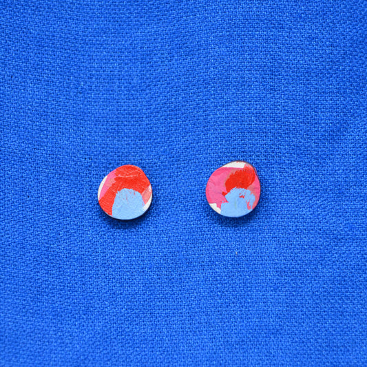 A pair of wooden round stud earrings with a colorful abstract pattern painted of red, pink, white and blue, displayed on a textured blue background.