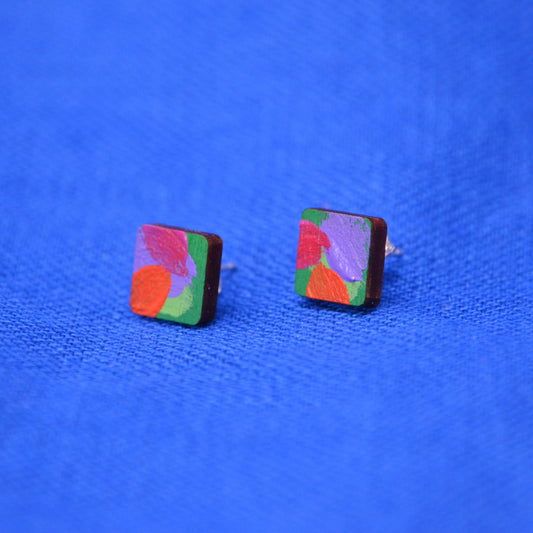 A pair of square stud earrings with colorful abstract painted designs on a dark wood base, displayed on a textured blue background.