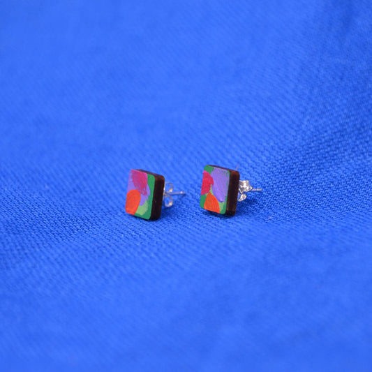 A pair of wooden square stud earrings with a pink, orange, green and purple painted abstract design, displayed on a textured blue background.