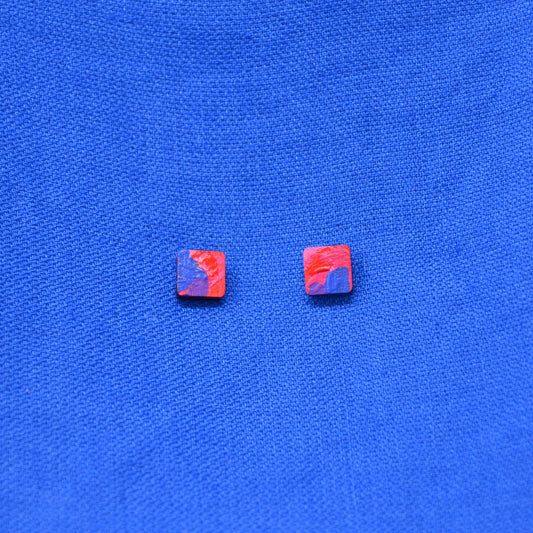 A pair of small wooden square stud earrings with a red, pink, and blue painted pattern, displayed on a textured blue fabric background.