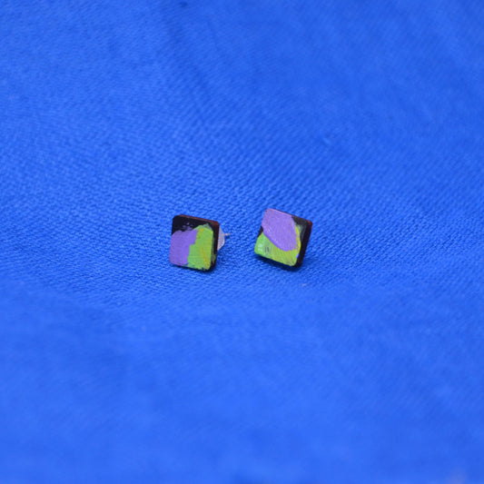 A pair of square stud earrings with a black base and an abstract paint design in purple and green, displayed on a textured blue background.