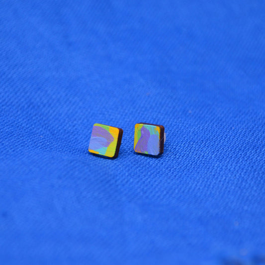 A pair of small wooden square stud earrings with a painted pattern of yellow, purple, and blue, displayed on a textured blue fabric background.