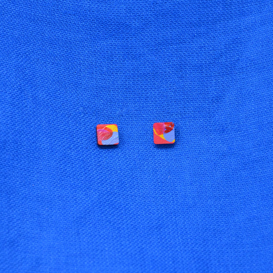 A pair of small wooden square stud earrings with a painted pattern of yellow, red, pink and blue, displayed on a textured blue fabric background.