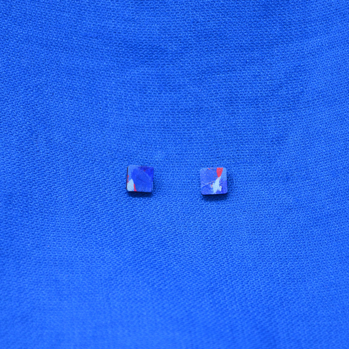 A pair of wooden square stud earrings with a painted blue and red pattern on a textured blue background.