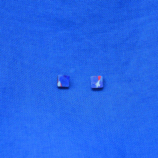 A pair of wooden square stud earrings with a painted blue and red pattern on a textured blue background.