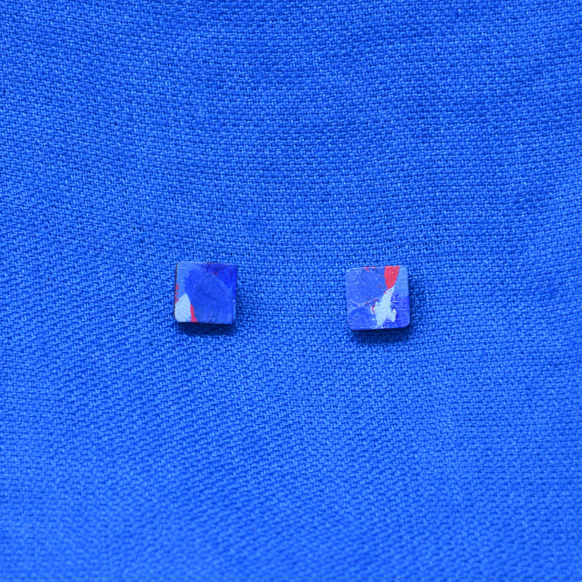 Two small square stud earrings with a marbled blue, purple, and red pattern, displayed on a textured blue fabric background.