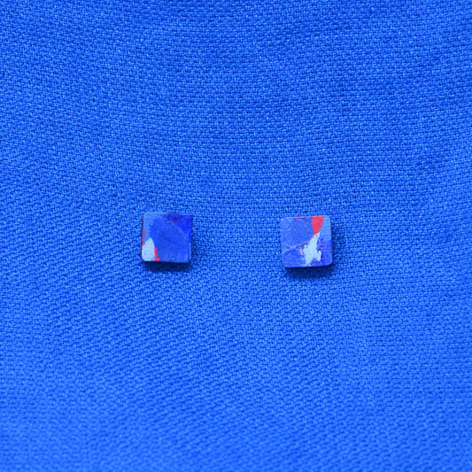 Two small square stud earrings with a marbled blue, purple, and red pattern, displayed on a textured blue fabric background.